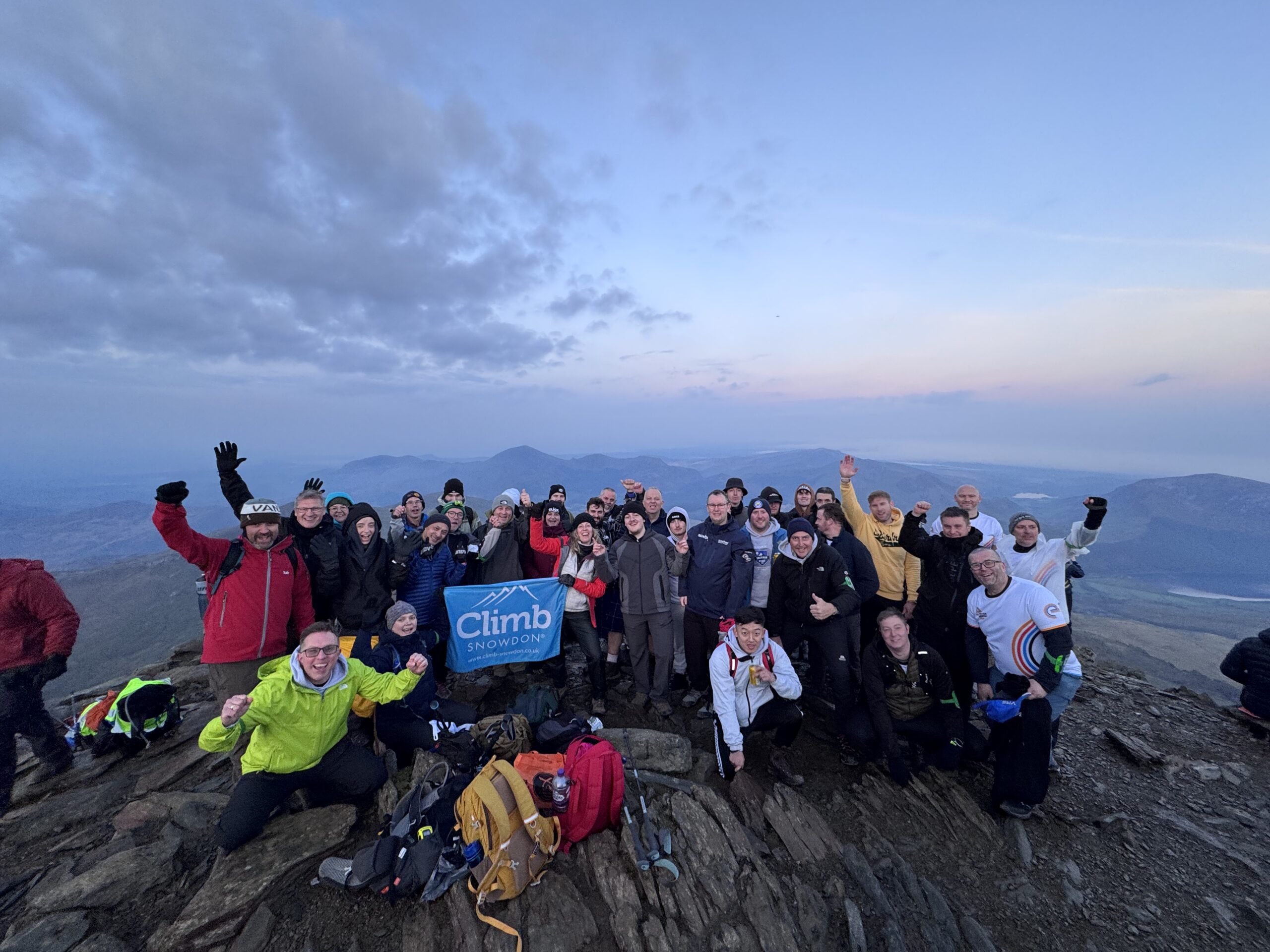 Large group of climbers smiling at the top of mount snowdon with sunrise behind them