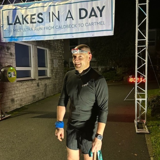 Philip Holt wearing running gear, standing in the dark at the Lakes in a Day finish line with a smile on his face